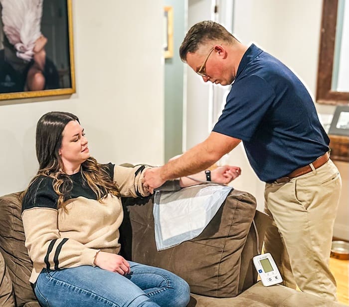 A nurse carefully wraps a blood pressure cuff around a woman's arm in preparation for a measurement, suggesting a healthcare check-up or monitoring scenario at home, possibly before administering IV hydration.